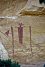 The 'Skeleton Shaman'. There is now a single snake and it is above the head of the main figure. There is a single hollow circle with no streamers above the main figure's head. The main figure has two distinct eyes with no pupils shown. The main figure and the 'monkey' like figure to the left  have holes in both their hands and feet. The 'monkey' figure and the snake have their respective tongues out and appear to be looking at each other. The 'ribs' of the main figure are clearly visible. The torso of the main figure is created with vertical striping that is not completely filled in. Significant??  The main figure has only four fingers on each hand and no elbow is indicated.  Note that both hands on the shaman are at or above shoulder level.