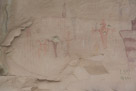 Moving on down the east wall we come to these figures. This is much more substantial. On close inspection here there is a lot to analyze.  First there are a number of colors present. Some of the images are quite faint. Does this indicate they are substantially older?? Or created using less permanent pigment.  The faint black figure in the upper center is tapered, has eyes and apparently horns.  Then there is that figure of the 14 vertical lines hanging from a horizontal line. Note the 6 vertical 'tic' marks between the orange and maroon tapered figures on the right edge. 
On the left is a faint red figure with a headdress and 6 descending vertical lines.  Then the orange figure with the tapered body and two antennae. Then there is the red Barrier Canyon figure that has suffered a rifle shot.  Despite this is apparent the this figure had at least one antenna, two purposeful hands and possible serpents suspended from each.  Definitely from his left shoulder. 
And yes, I see the graffiti.
