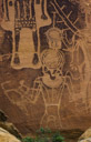 Sort of hard to sort things out with this character. There appears to be newer and older glyphs interferring with this figure. 
An elaborate breast plate is clearly indicated on a Fremone trapazoid torso. I believe there is a headdress with  three feathers? indicated on each side. The face is covered by a mask. The torso is decorated with three rows of beads? highlighting the trapazoid shape. Arms and legs are indicated but not developed. A single line indicates a simple belt.