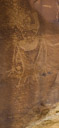 This figure exhibits the trapazoid torso of the Fremont. There is a fully pecked out breast plate. Eyes and mouth are indicated but no ears or ear treatment. There may be a nose indicated as well. The eyes appear to  have tear streaks to the sides. A wide decorated belt is indicated. The headdress, possibly matching the belt is accented with three feathers? on each side. Arms are drawn and hands are fully pecked. Legs are indicated. Both arms and legs appear to be done for completeness. The torso is not decorated.